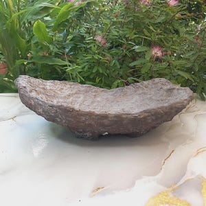 Puede incluir: Un cuenco oblongo hecho a mano, con aspecto de piedra y superficie rugosa y texturizada. El cuenco es de color marrón apagado y descansa sobre tres pequeños pies redondeados. El fondo presenta un exuberante follaje verde y flores rosas.