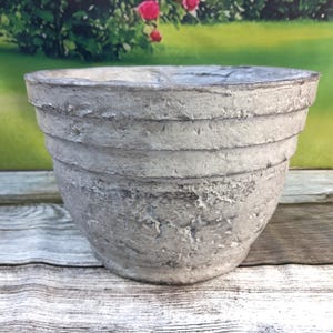 May include: A weathered gray concrete planter with a textured surface and a tiered design. The planter has a wide opening and is suitable for outdoor use. The background shows a garden scene with green grass and red roses.