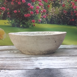 Op de afbeelding: Een lichtgrijze, ovaalvormige plantenbak met een ruw, getextureerd oppervlak. De plantenbak heeft een brede opening en een ondiepe diepte. De achtergrond toont een tuin met roze rozen en groen gras. Geschikt voor kleine planten en vetplanten.