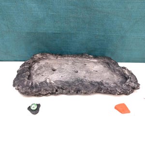 Peut inclure: Un plateau rectangulaire avec une bordure texturée gris foncé rappelant la roche et une surface intérieure plate gris clair comportant plusieurs petites indentations circulaires espacées. Un anneau vert et une pièce triangulaire orange.