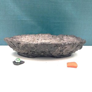 Pode incluir: Uma tigela de cerâmica ovalada, texturizada, cinza escuro, com uma borda áspera e irregular. Uma pequena bolinha de gude de vidro verde está sobre uma pedra cinza escura, e um pequeno pedaço laranja está ao lado da tigela. A tigela está em uma superfície branca.