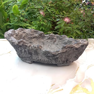 Op de afbeelding: Een donkergrijze, langwerpige plantenbak met een ruw, getextureerd oppervlak dat het uiterlijk van natuursteen of schors nabootst. De bak heeft een onregelmatige, organische rand en rust op kleine, geïntegreerde voetjes aan de onderkant.