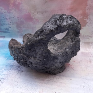 May include: A dark gray, textured, rock-like sculpture with an open center. The object has a rough, uneven surface and appears to be hand-crafted. The sculpture is set against a colorful, abstract background.