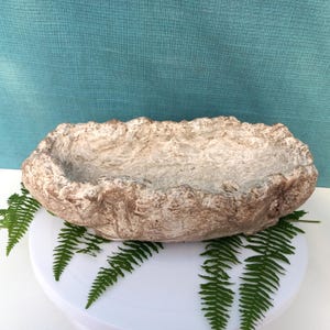 Op de afbeelding: Een beige, ovaalvormige plantenbak met een textuur die op steen lijkt. De plantenbak heeft een ruwe, ongelijke rand en wordt getoond op een wit oppervlak met groene varenbladeren. De achtergrond is effen blauwgroen.