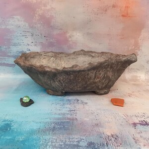 Peut inclure: Un pot de fleurs ovale marron texturé avec une surface rugueuse et inégale. Le pot repose sur de petits pieds et possède un intérieur clair. Une petite bille verte et un éclat orange sont à côté du pot.