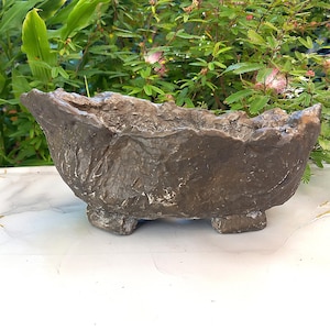 Può includere: Un vaso decorativo marrone strutturato con una forma organica ruvida. Il vaso poggia su tre piccoli piedi e ha una parte superiore ampia e aperta. Lo sfondo presenta un fogliame verde.