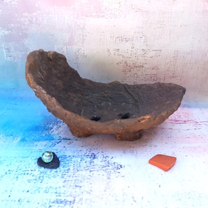 Peut inclure: Un bol en céramique marron à la texture organique, doté d'un bord surélevé irrégulier et de trois petits pieds. La base intérieure présente deux trous de drainage circulaires. Une bille en verre transparent sur une pierre sombre et un fragment de tuile orange sont posés à proximité.