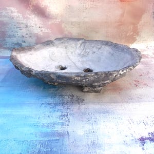 Può includere: Un vaso ovale in ceramica per bonsai con un esterno ruvido e strutturato nei toni del grigio scuro e dell'argento. L'interno presenta una finitura liscia grigio chiaro con due fori di drenaggio sul fondo. Il vaso poggia su piccoli piedini.
