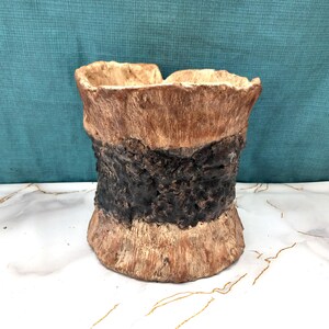 May include: A textured, handcrafted planter with a rustic appearance. The planter has a layered design with a light brown top, a dark brown band, and a lighter brown base. The open top suggests it is designed for plants or decorative items.