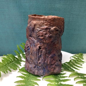 Può includere: Un vaso in ceramica fatto a mano, strutturato, con un aspetto rustico simile alla corteccia. Il vaso è principalmente marrone con sfumature blu e poggia su una superficie bianca con foglie di felce verdi. Il vaso cilindrico ha una superficie ruvida e irregolare.