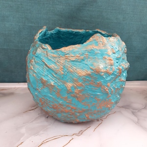 Puede incluir: Una maceta redonda, texturizada, pintada de color turquesa y dorado. El exterior tiene una apariencia rugosa y orgánica, con el color base turquesa acentuado por reflejos dorados. El interior es turquesa.
