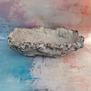 Può includere: Una fioriera oblunga con un aspetto strutturato simile alla pietra. La fioriera è principalmente bianco sporco con accenti grigio scuro lungo i bordi, creando un'estetica naturale e invecchiata. L'interno è liscio.