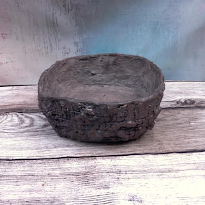 Puede incluir: Un cuenco ovalado de color gris oscuro con un exterior rugoso y texturizado que imita la apariencia de la corteza de un árbol. El interior del cuenco es liso y cóncavo, presentando una cuenca profunda con un acabado gris oscuro uniforme.