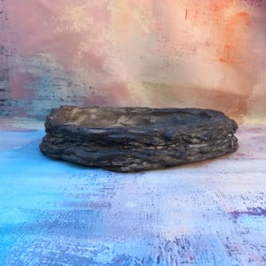May include: A dark grey, textured, rectangular planter with a rough, bark-like appearance. The planter has a shallow, open design, suitable for small plants or succulents. The interior is a lighter grey, contrasting with the darker exterior. The background is a colourful blend of pink, orange, and blue.