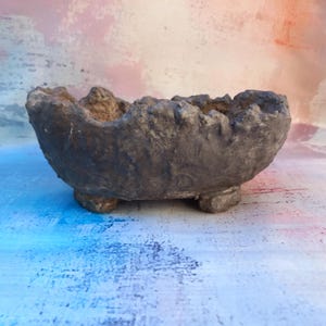 May include: A gray, textured concrete planter with a rough, uneven rim. The small, round feet elevate the bowl-shaped container. The planter is suitable for small plants and succulents.