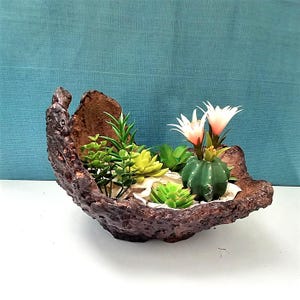 Op de afbeelding: Een bruine, getextureerde, komvormige plantenbak gevuld met kunstmatige vetplanten en een kleine cactus met witte en roze bloemen. De plantenbak staat op een wit oppervlak tegen een turquoise achtergrond.