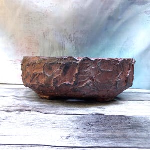 Pode incluir: Um vaso de cerâmica redondo e de perfil baixo com um exterior rugoso e texturizado em tons de castanho-avermelhado profundo e cinzento escuro. O recipiente apresenta um rebordo espesso e irregular e pequenos pés integrados na base.