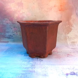 Può includere: Un vaso di fiori ottagonale in ceramica marrone con un bordo leggermente svasato. Il vaso ha una superficie strutturata e poggia su quattro piedini. Lo sfondo è una miscela di blu, rosa e bianco.