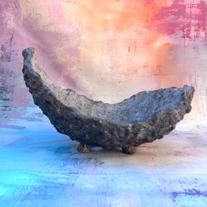 Puede incluir: Un cuenco decorativo gris texturizado y hecho a mano, con forma curva y tres patas pequeñas. El cuenco tiene un aspecto rugoso y orgánico y está colocado sobre un fondo colorido y abstracto con tonos de rosa, azul y naranja.