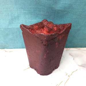 Può includere: Un vaso quadrato, di colore bordeaux scuro con una superficie strutturata. L'interno è di un rosso vibrante. Il vaso ha un bordo unico e irregolare ed è adatto a piccole piante o succulente. Lo sfondo è un tessuto color verde acqua.