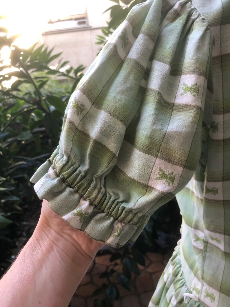 Op de afbeelding: Een groene en witte geruite jurk met een geplooide mouwdetail. De jurk heeft een wit geborduurd patroon over de hele oppervlakte.