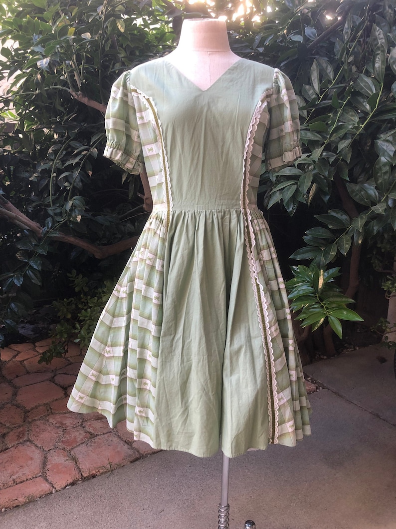 Op de afbeelding: Een groene vintage jurk met een wit en groen geruit patroon op de rok en mouwen. De jurk heeft een V-hals en een getailleerd bovenlijf. De jurk staat op een mannequin.