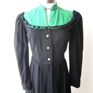 Könnte beinhalten: Ein schwarzes, langärmliges Kleid mit einem grünen, gekräuselten Kragen und silbernen Knöpfen. Das Kleid hat ein tailliertes Oberteil und einen fließenden Rock.