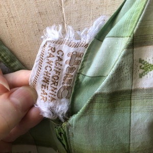 Op de afbeelding: Een groene en witte geruite jurk met een rits en een label met de tekst "California Ranchwear" in bruine letters.