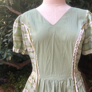 Op de afbeelding: Een groene vintage jurk met witte kant en een gestreept patroon op de mouwen. De jurk heeft een V-hals en een geplooide rok.