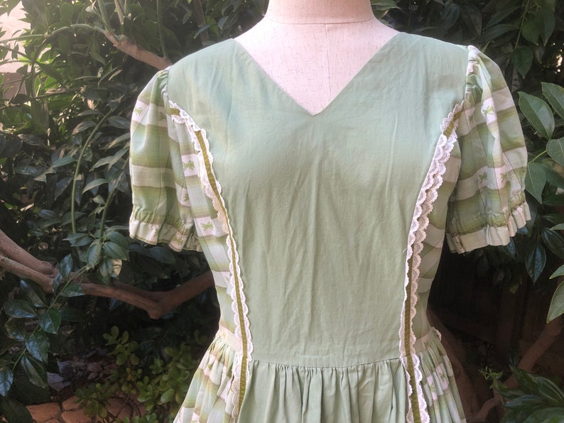 Op de afbeelding: Een groene vintage jurk met witte kant en een gestreept patroon op de mouwen. De jurk heeft een V-hals en een geplooide rok.