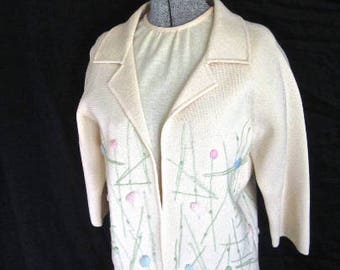May include: A white cardigan with a delicate floral design in shades of pink, blue, and green. The cardigan has a long sleeve and a relaxed fit.