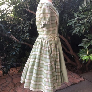 Op de afbeelding: Een groene en witte gingham jurk met een strikprint. De jurk heeft een getailleerd bovenlijf en een volle rok. De mouwen zijn kort en gepoft.
