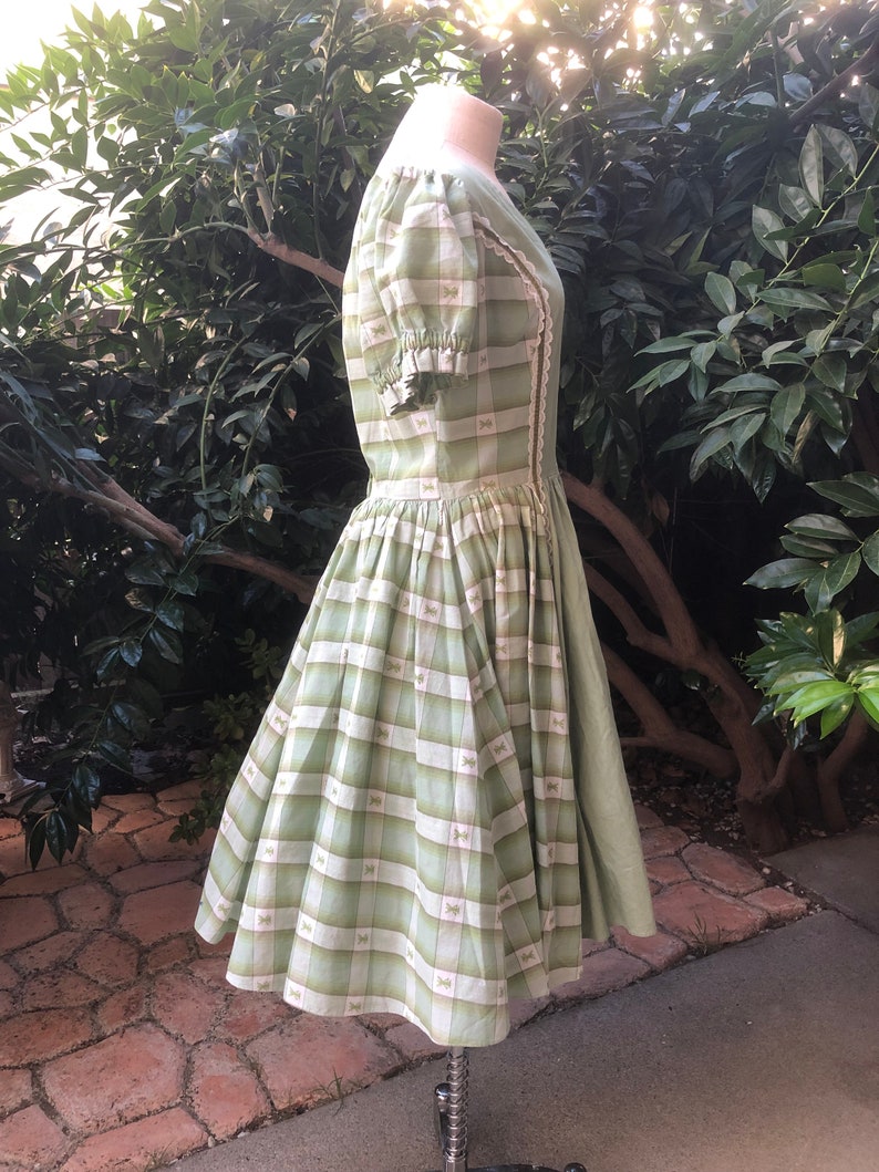 Op de afbeelding: Een groene en witte gingham jurk met een strikprint. De jurk heeft een getailleerd bovenlijf en een volle rok. De mouwen zijn kort en gepoft.