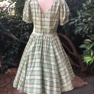 Op de afbeelding: Een groene en witte gingham jurk met een strikprint. De jurk heeft een v-hals, korte pofmouwen en een volle rok.
