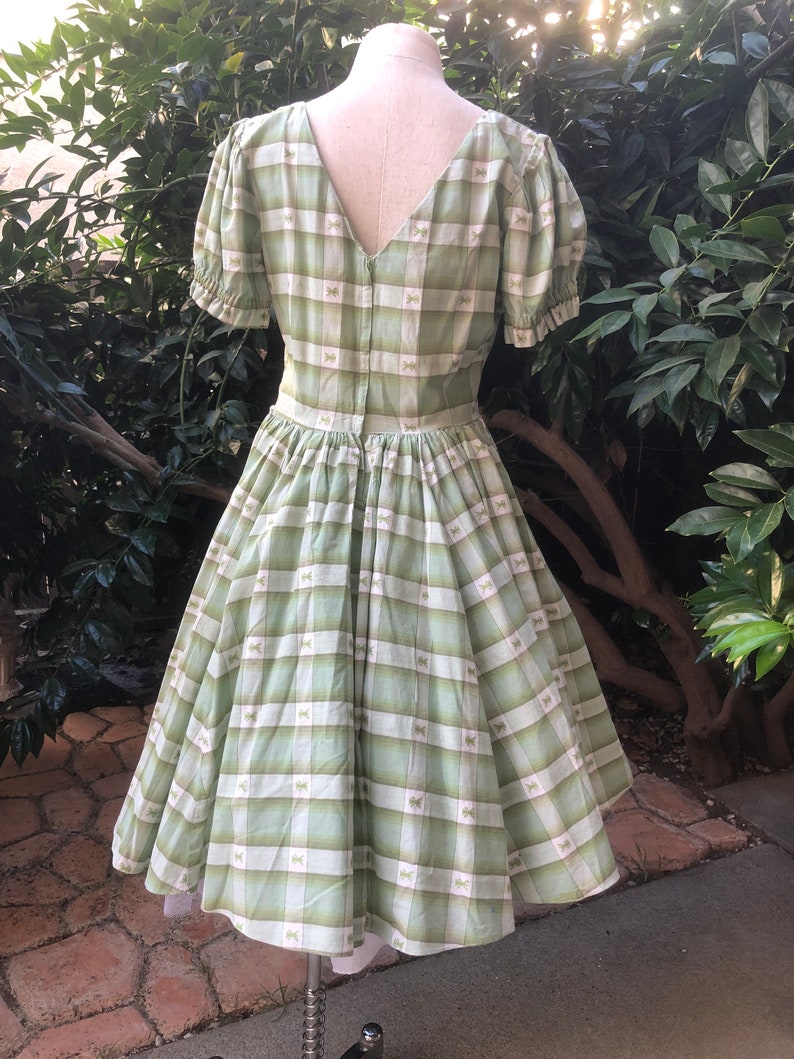 Op de afbeelding: Een groene en witte gingham jurk met een strikprint. De jurk heeft een v-hals, korte pofmouwen en een volle rok.