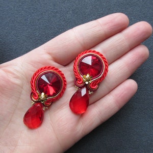 Può includere: Un paio di orecchini rossi importanti. Ogni orecchino presenta un grande cristallo rosso sfaccettato al centro, circondato da un design intrecciato rosso e oro. Un cristallo rosso a forma di goccia pende sotto, con piccoli accenti dorati.