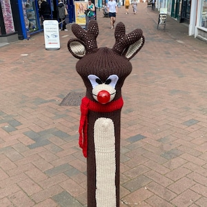 May include: A knitted reindeer sculpture on a street post. The reindeer is brown and white with a red nose and a red scarf.
