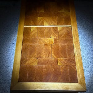 May include: A rectangular wooden cutting board with a light brown frame and a dark brown center made of small, square pieces of wood.