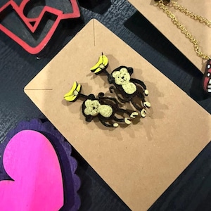 Puede incluir: Pendientes con forma de mono con detalles de plátanos amarillos, expuestos sobre una tarjeta marrón. Los pendientes presentan una combinación de colores negro y dorado, con un diseño lúdico. También se ven otros elementos decorativos, como un recorte de "LOVE" rojo y un corazón rosa.