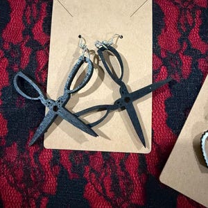 May include: Black scissor-shaped earrings with silver-colored ear wires are displayed on a brown card. The earrings are made of a dark material and are set against a red and black patterned background.