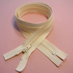 Puede incluir: Cremallera color crema, enrollada y colocada sobre una superficie rosa. La cremallera presenta un cursor y dientes metálicos, con la cinta de tela que muestra un sutil patrón de espiga. Para proyectos de costura.