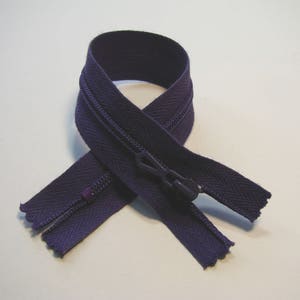 May include: A dark purple zipper with a matching pull tab is displayed against a white background. The zipper is made of a woven material and has a closed design. The zipper is a common sewing supply.