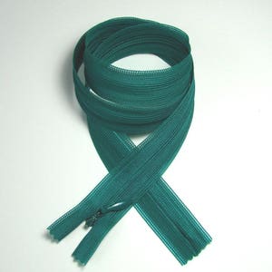 May include: A teal-colored zipper, coiled and crossed, against a white background. The zipper's teeth are visible, and the pull tab is in the closed position. The fabric has a fine, woven texture.