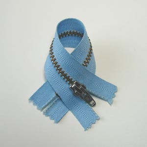 May include: A light blue zipper with a silver pull tab and bronze-colored teeth. The zipper is looped and crossed over itself, showcasing its construction and color. The zipper is a common component in clothing and accessories.
