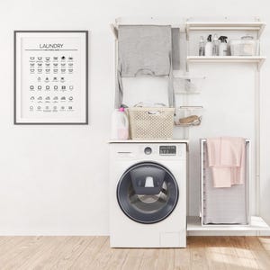May include: A white washing machine with a dark blue door is positioned below a laundry organization system. The system includes shelves, a hanging rod with a gray shirt, and a framed "LAUNDRY" care symbol guide.