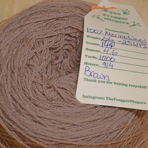 Może przedstawiać: Motka brązowej przędzy z 100% wełny merino, 2-nitkowa, z etykietą z napisem "The Frogger Niagara" i szczeg&oacute;łami takimi jak waga (1244 gramy lub 4,6 uncji), długość (1000 jard&oacute;w) i metraż (914 metr&oacute;w).