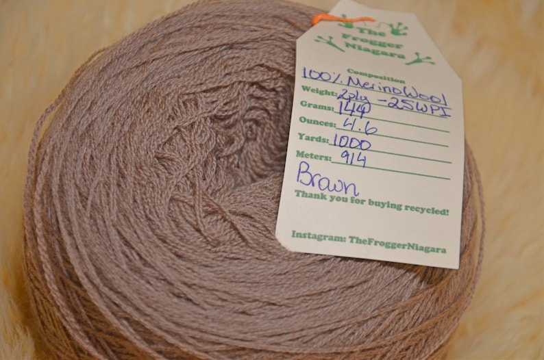 Może przedstawiać: Motka brązowej przędzy z 100% wełny merino, 2-nitkowa, z etykietą z napisem "The Frogger Niagara" i szczeg&oacute;łami takimi jak waga (1244 gramy lub 4,6 uncji), długość (1000 jard&oacute;w) i metraż (914 metr&oacute;w).