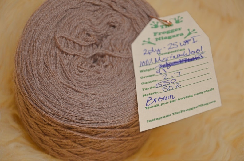 Może przedstawiać: Motka brązowej przędzy wełnianej merino 2-nitkowej z etykietą z napisem "Frogger Niagara 2ply. 25 WPI 100% Wełna Merino Skład Waga: 27g Gramy: 2,7 Uncje: 550 Yardy: 502 Metry: Brązowy Dziękujemy za zakup recyklingu! Instagram TheFrogger Niagara"