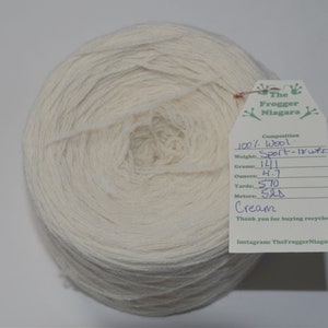 May include: A skein of cream-colored wool yarn, labeled "The Frogger Niagara" with details about the yarn's composition, weight, and yardage. The label reads "100% Wool, Sport - 18 wpi, 141 grams, 4.7 ounces, 570 yards, 520 meters, Cream".