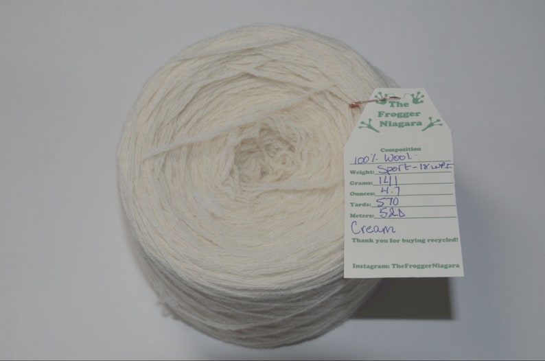 May include: A skein of cream-colored wool yarn, labeled "The Frogger Niagara" with details about the yarn's composition, weight, and yardage. The label reads "100% Wool, Sport - 18 wpi, 141 grams, 4.7 ounces, 570 yards, 520 meters, Cream".